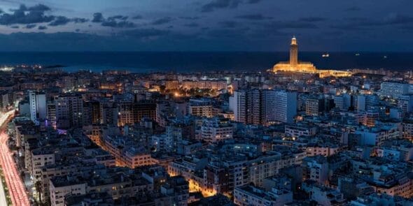 Vue aérienne d'un paysage urbain au crépuscule avec des bâtiments illuminés et une tour éclairée proéminente près du littoral, où l'horizon est orné de lieux populaires comme le Restaurant à Casablanca.