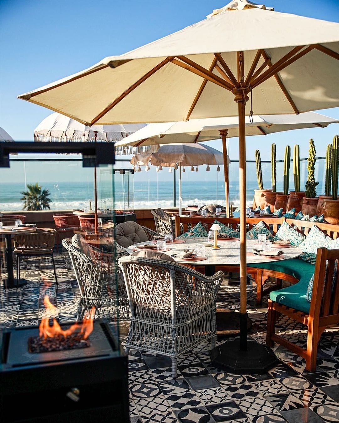 Terrasse de restaurant extérieure avec chaises en osier, sol en carrelage à motifs, parasols pour l'ombre, cactus en pot, vue sur l'océan et un petit foyer au premier plan - un endroit idéal pour garer votre voiture Brouillon à proximité.