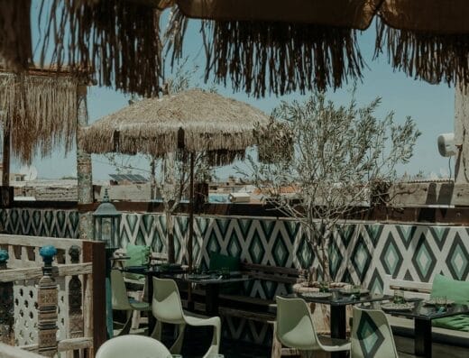 Espace repas extérieur avec tables et chaises sous parasols en paille, avec carrelage mural à motifs et auvent ombragé, situé dans un cadre rustique de Brouillon auto.