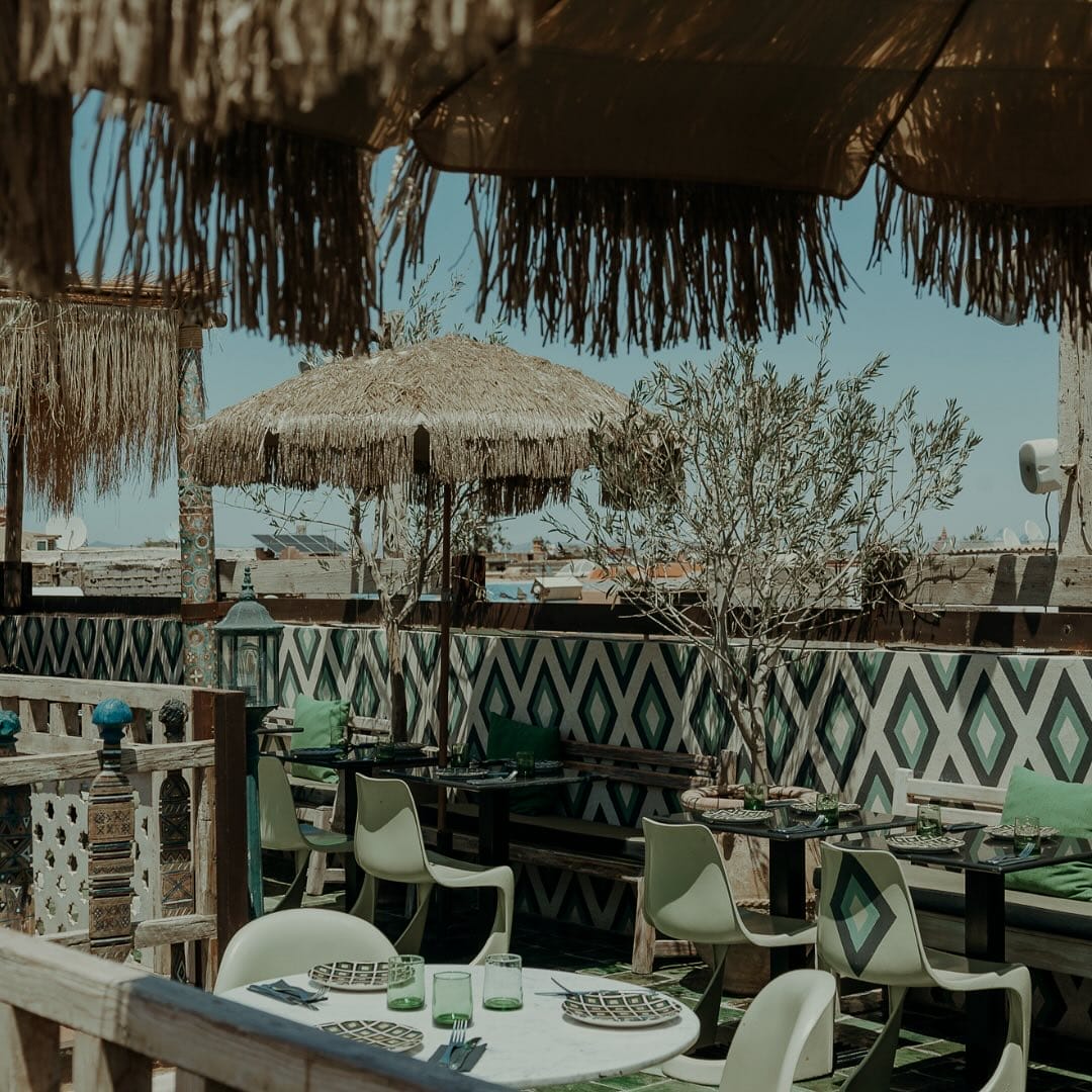 Espace repas extérieur avec tables et chaises sous parasols en paille, avec carrelage mural à motifs et auvent ombragé, situé dans un cadre rustique de Brouillon auto.