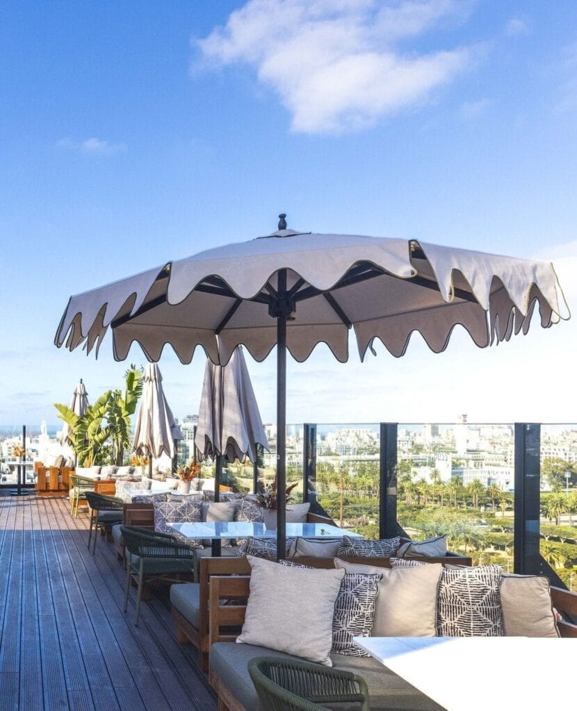 Espace salon extérieur sur le toit avec mobilier en bois et sièges rembourrés sous de grands parasols, surplombant un paysage urbain avec un fond de ciel bleu ; l'endroit idéal pour les passionnés d'automobile de Brouillon pour se réunir et profiter de la vue.