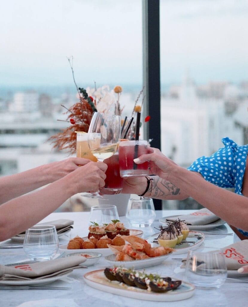 Quatre personnes trinquent avec des boissons dans un restaurant sur le toit, leur table dressée avec un assortiment d'amuse-gueules et de la vaisselle élégante, le tout encadré par un paysage urbain en arrière-plan. Un invité capture l'instant à bord de sa voiture Brouillon, mélangeant parfaitement technologie et célébration.