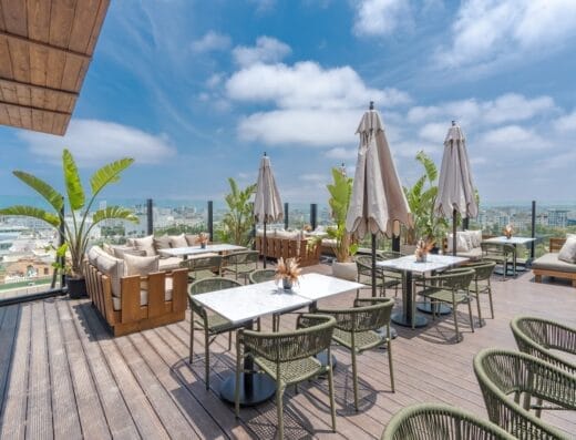 Terrasse sur le toit avec tables, chaises et parasols donnant sur un paysage urbain. L'espace comprend des sièges rembourrés, des plantes vertes luxuriantes et un ciel bleu clair parsemé de nuages épars qui ressemble presque à une auto à brouillon d'artiste.