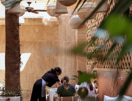 Un serveur s'occupe des clients sur la terrasse extérieure du restaurant Brouillon, ornée de meubles en bois, de lanternes suspendues et d'un palmier majestueux se balançant doucement dans la brise.