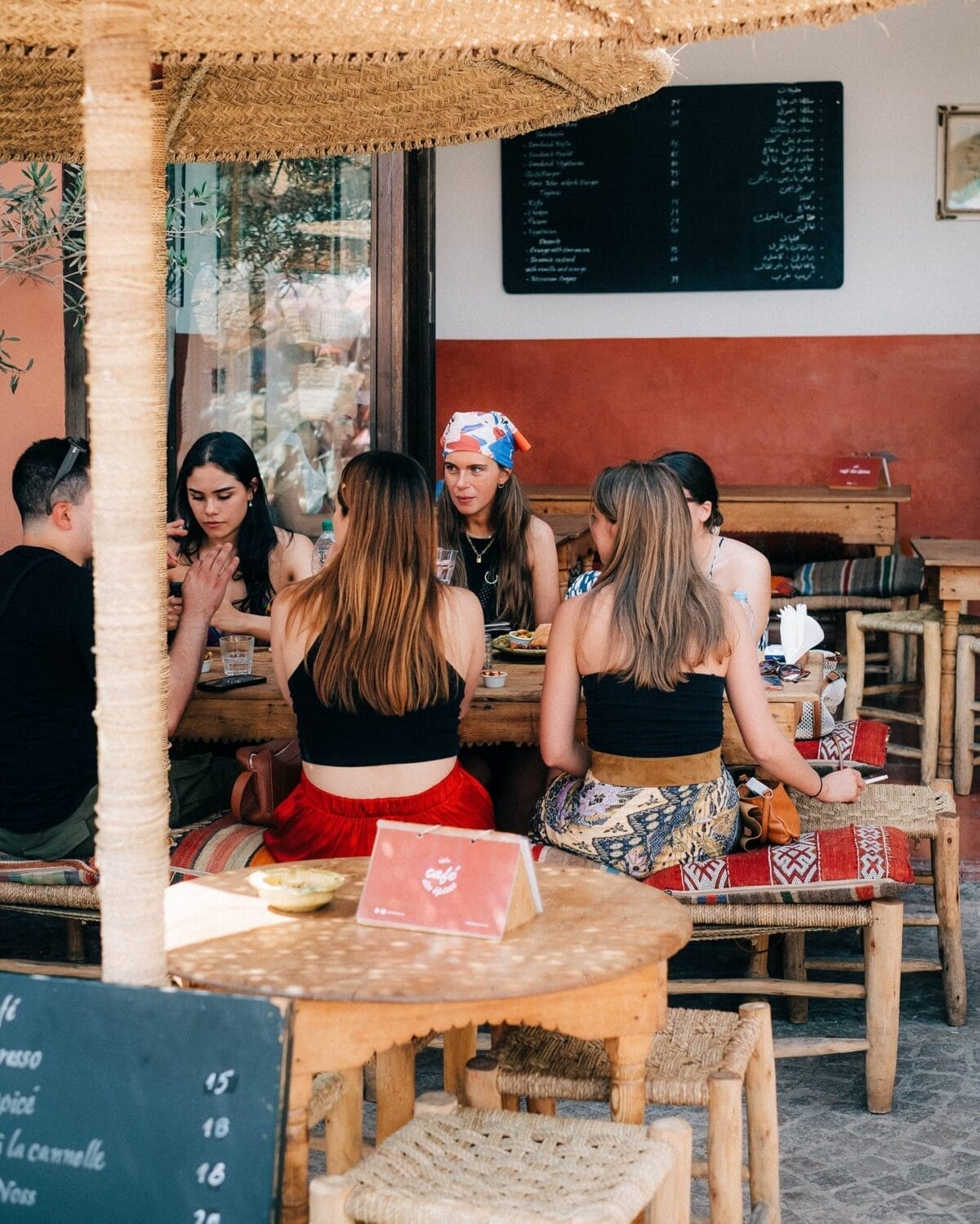 Un groupe de personnes sont assises autour d'une table en bois dans un café en plein air, engagées dans une conversation animée. Le café, nommé Brouillon Auto, arbore un panneau de menu en arrière-plan et est décoré de chaises tressées et de meubles rustiques.