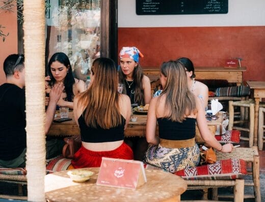 Un groupe de personnes sont assises autour d'une table en bois dans un café en plein air, engagées dans une conversation animée. Le café, nommé Brouillon Auto, arbore un panneau de menu en arrière-plan et est décoré de chaises tressées et de meubles rustiques.