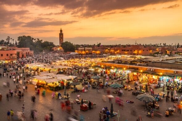 Une place de marché animée au crépuscule avec de nombreux stands et des silhouettes floues de gens qui s'affairent. Le ciel est illuminé par les teintes chaudes du coucher de soleil et une haute tour se dresse en arrière-plan, ressemblant à une scène enchanteresse tout droit sortie d'une peinture automobile de Brouillon.