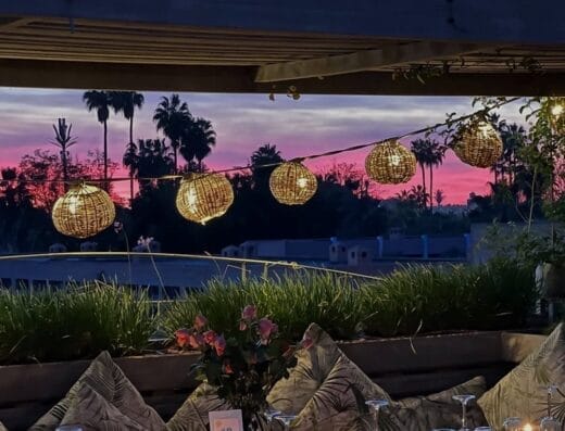 Espace repas extérieur au coucher du soleil avec pergola, lanternes suspendues et décorations florales, créant une atmosphère pittoresque de brouillon auto. Le ciel est vibrant de nuances de rose et de violet.