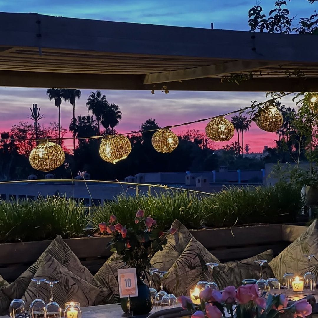 Espace repas extérieur au coucher du soleil avec pergola, lanternes suspendues et décorations florales, créant une atmosphère pittoresque de brouillon auto. Le ciel est vibrant de nuances de rose et de violet.