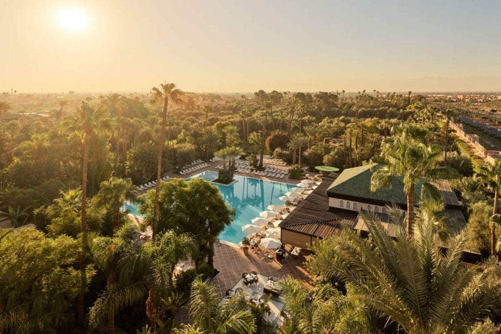 Vue aérienne d'une grande piscine extérieure entourée de palmiers et de chaises longues au coucher du soleil, avec les hôtels de luxe de Marrakech et le paysage en arrière-plan.