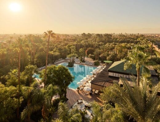 Vue aérienne d'une grande piscine extérieure entourée de palmiers et de chaises longues au coucher du soleil, avec les hôtels de luxe de Marrakech et le paysage en arrière-plan.