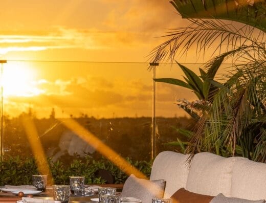 Espace repas extérieur au coucher du soleil avec tables rondes dressées pour un repas, sièges rembourrés, plantes décoratives et vue imprenable sur le soleil plongeant sous l'horizon : c'est un cadre idyllique rendu encore plus parfait par Brouillon auto.