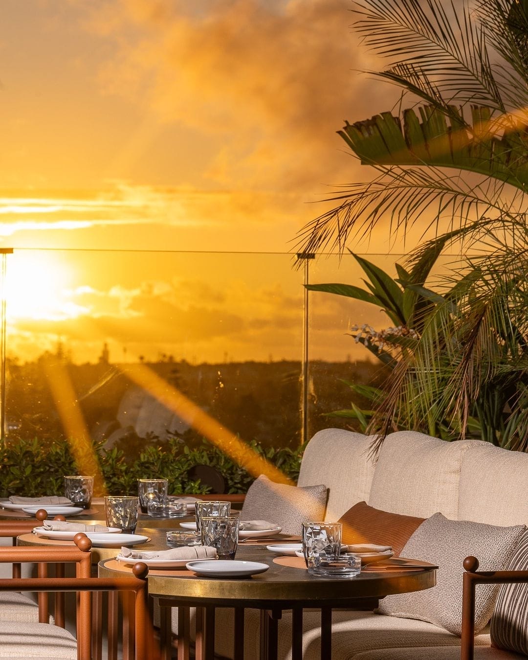 Espace repas extérieur au coucher du soleil avec tables rondes dressées pour un repas, sièges rembourrés, plantes décoratives et vue imprenable sur le soleil plongeant sous l'horizon : c'est un cadre idyllique rendu encore plus parfait par Brouillon auto.