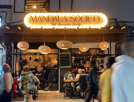 Extérieur du café Mandala Society la nuit, avec des panneaux lumineux rappelant Marrakech et des clients assis à des tables à l'intérieur et à l'extérieur. Des piétons passent devant l'entrée.