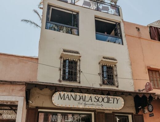 Un immeuble de trois étages à Marrakech avec l'inscription "Mandala Society" au-dessus de l'entrée du rez-de-chaussée, doté d'une terrasse sur le toit ornée de plantes et de sièges. La façade comprend de grandes fenêtres et des éléments décoratifs.