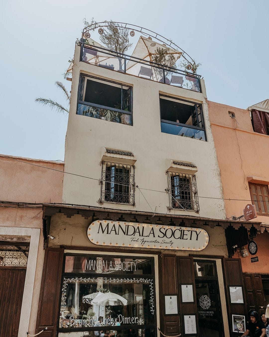 Un immeuble de trois étages à Marrakech avec l'inscription "Mandala Society" au-dessus de l'entrée du rez-de-chaussée, doté d'une terrasse sur le toit ornée de plantes et de sièges. La façade comprend de grandes fenêtres et des éléments décoratifs.