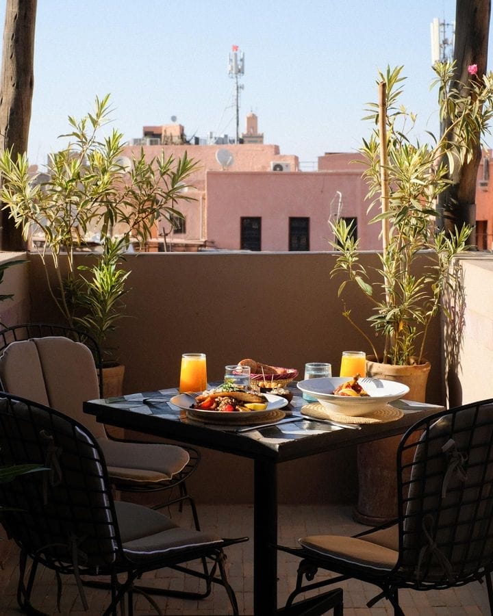 Un toit-terrasse avec une table dressée pour un repas, comprenant des assiettes de nourriture et de boissons, surplombe les bâtiments éloignés et les plantes en pot sous un ciel bleu clair. L’ambiance est parfaite pour capturer des moments spontanés semblables à un brouillon auto.