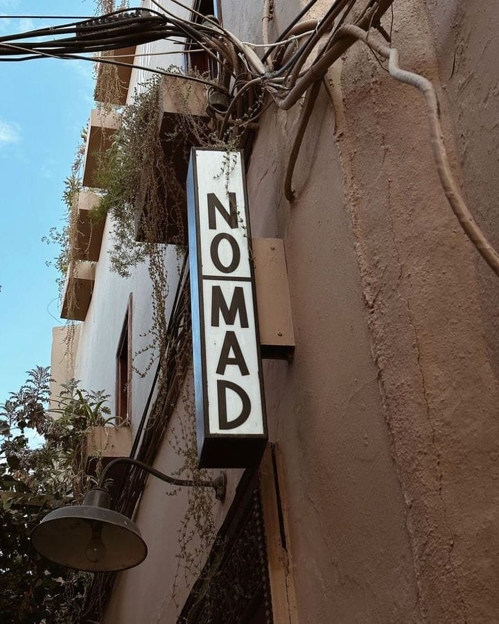 Un panneau vertical indiquant « NOMAD » est fixé à l'extérieur d'un bâtiment beige, avec des fils électriques et des plantes visibles en arrière-plan, lui conférant un charme automobile éclectique à Brouillon.