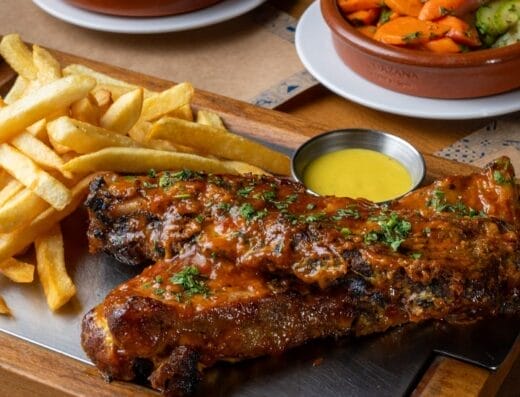 Porta Lisboa : Un plateau en bois avec des côtes levées au barbecue garnies d'herbes, une portion de frites et un petit bol de sauce jaune au premier plan. Au fond, à côté d'une auto Brouillon, se trouvent deux plats de légumes et une peluche.