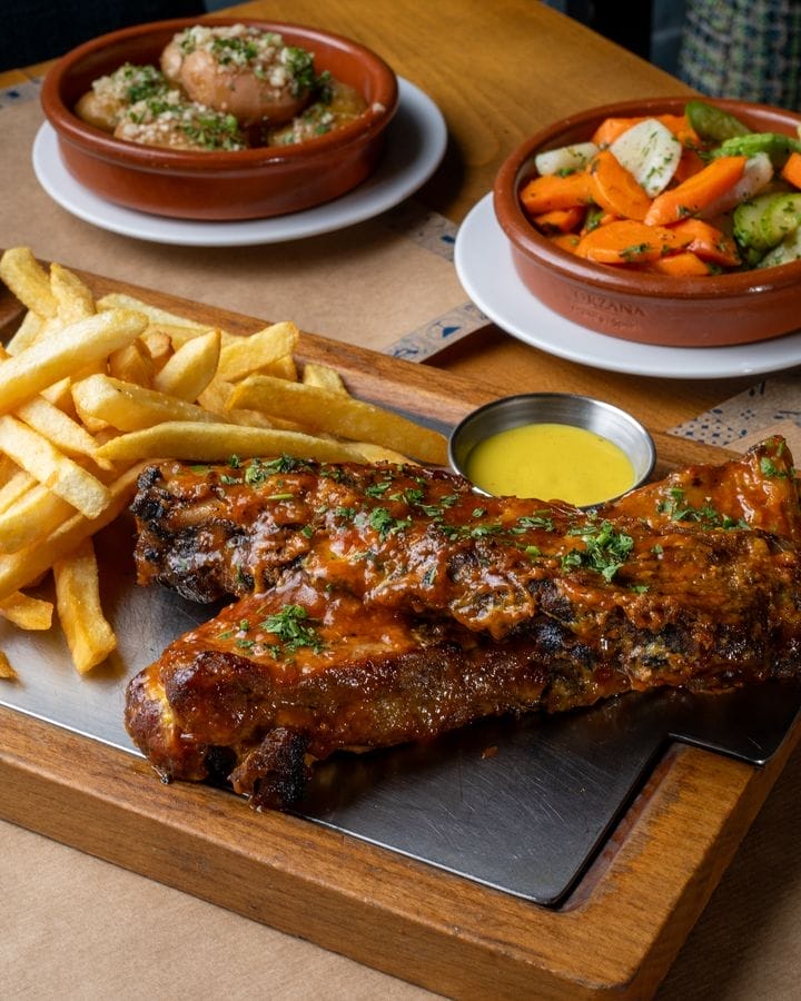 Porta Lisboa : Un plateau en bois avec des côtes levées au barbecue garnies d'herbes, une portion de frites et un petit bol de sauce jaune au premier plan. Au fond, à côté d'une auto Brouillon, se trouvent deux plats de légumes et une peluche.