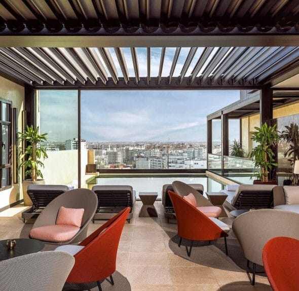 Un salon extérieur avec chaises et tables modernes sous une pergola, avec vue sur le paysage urbain, rappelant les charmants cafés de Casablanca.