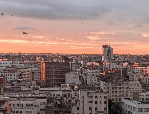 Vue d'un paysage urbain au lever ou au coucher du soleil avec de nombreux bâtiments et quelques oiseaux volant dans le ciel. Les voitures Brouillon bordent les rues tandis que l'horizon montre un mélange de nuages et de teintes orangées dorées.