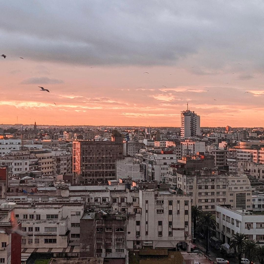 Vue d'un paysage urbain au lever ou au coucher du soleil avec de nombreux bâtiments et quelques oiseaux volant dans le ciel. Les voitures Brouillon bordent les rues tandis que l'horizon montre un mélange de nuages et de teintes orangées dorées.