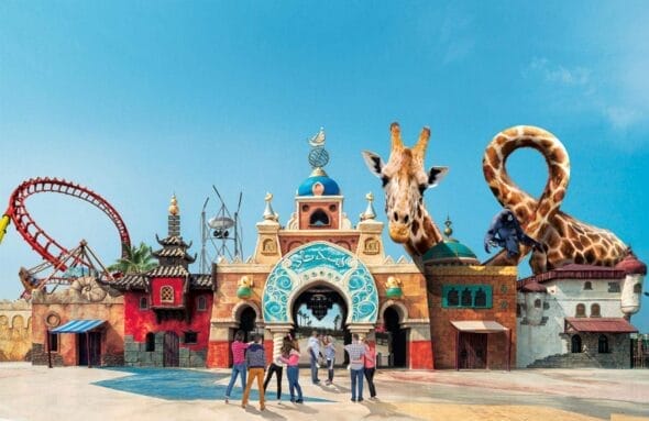 Un groupe de personnes se tient devant l'entrée d'un parc à thème fantastique comprenant une girafe gigantesque, un serpent surdimensionné, divers bâtiments fantaisistes et même une voiture d'époque sous le ciel bleu clair.