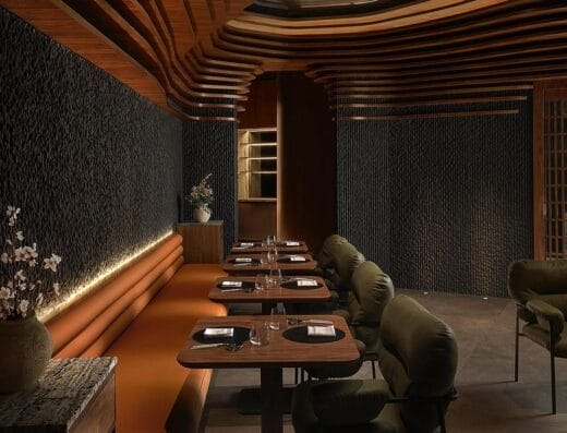 Une salle à manger de restaurant moderne avec des murs texturés sombres, un plafond en bois en forme de vague, une banquette marron à gauche, des chaises rembourrées vertes à droite et des tables garnies de verrerie et de serviettes. Le mobilier soigneusement choisi crée une atmosphère accueillante pour les convives.