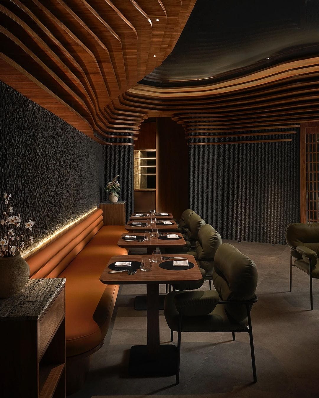 Une salle à manger de restaurant moderne avec des murs texturés sombres, un plafond en bois en forme de vague, une banquette marron à gauche, des chaises rembourrées vertes à droite et des tables garnies de verrerie et de serviettes. Le mobilier soigneusement choisi crée une atmosphère accueillante pour les convives.