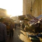 Une scène de marché en plein air ensoleillée avec des gens se promenant parmi les étals vendant divers produits, illuminés par la lumière dorée du soleil couchant, évoque le charme animé de la médina de Fès.