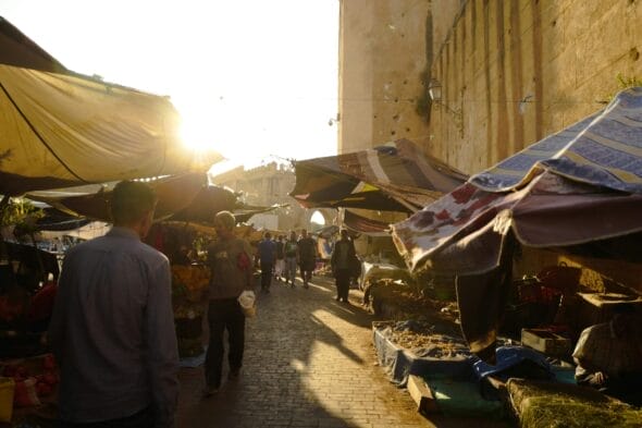 Une scène de marché en plein air ensoleillée avec des gens se promenant parmi les étals vendant divers produits, illuminés par la lumière dorée du soleil couchant, évoque le charme animé de la médina de Fès.