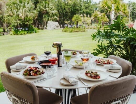 Une table ronde extérieure est dressée avec des assiettes de salades, diverses boissons et des serviettes, rappelant une expérience culinaire de Robuchon, sur fond de jardin verdoyant. Quatre chaises beiges entourent la table dans ce cadre de club-house chaleureux.