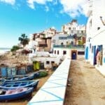 Un village côtier avec des bateaux colorés sur un rivage sablonneux, des bâtiments blanchis à la chaux bordant une passerelle et un ciel bleu clair au-dessus évoque le charme du Maroc.