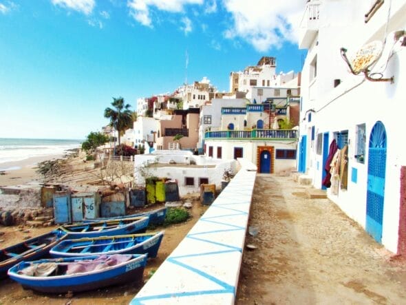 Un village côtier avec des bateaux colorés sur un rivage sablonneux, des bâtiments blanchis à la chaux bordant une passerelle et un ciel bleu clair au-dessus évoque le charme du Maroc.