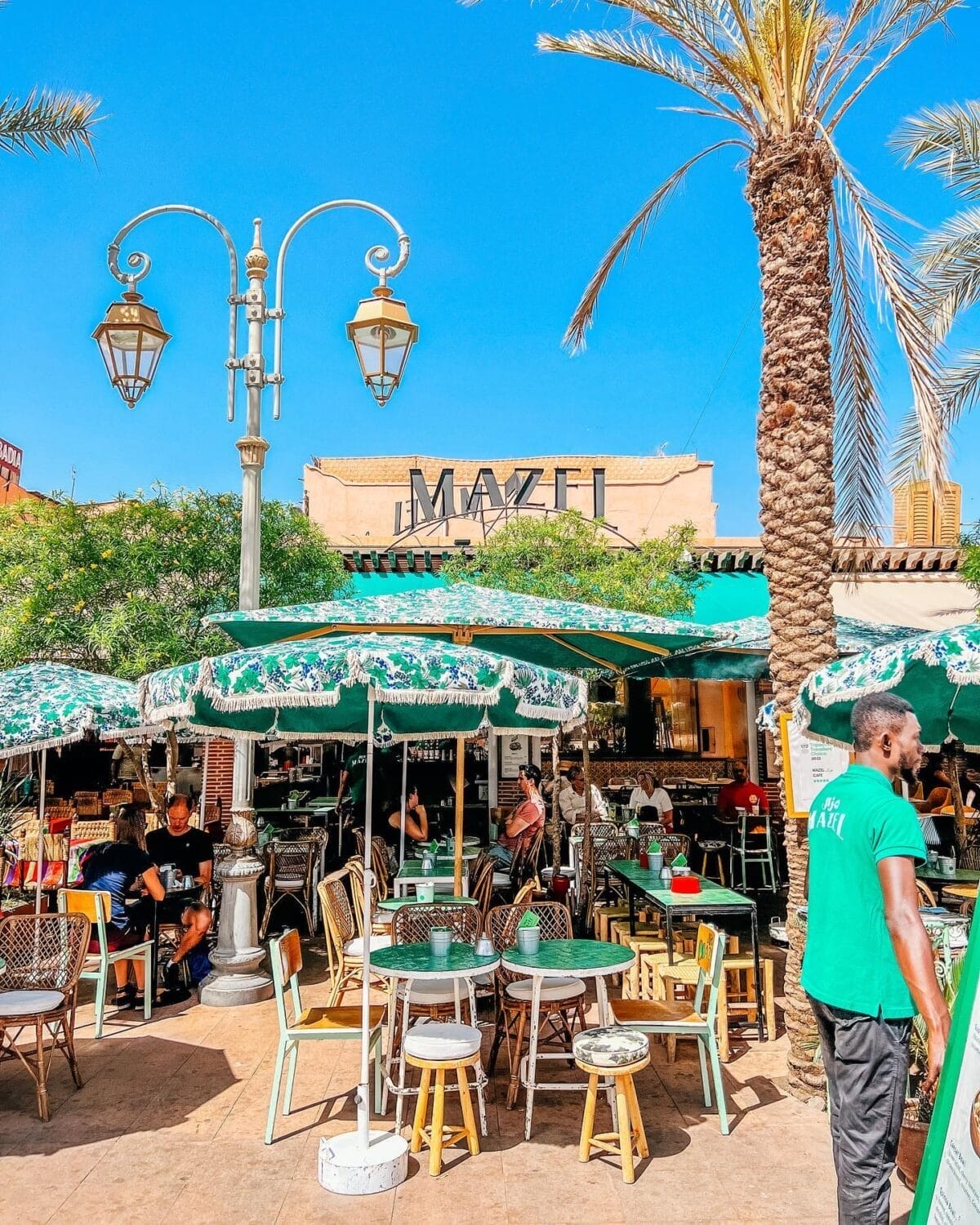 Le café en plein air, orné de parasols verts, de tables et de chaises, met en valeur le panneau « MAZEL » en arrière-plan. Un membre du personnel en chemise verte se tient à proximité. Des palmiers et des lampadaires vintage ajoutent du charme sous un ciel bleu clair.