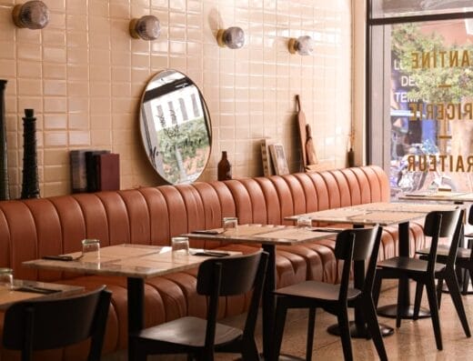 Un intérieur de café moderne avec des banquettes en cuir beige le long d'un mur carrelé avec des miroirs ronds, des chaises noires, des petites tables en bois et une grande fenêtre avec lumière naturelle. L'ambiance est subtilement rehaussée par des touches de raffinement Mouton Noir.