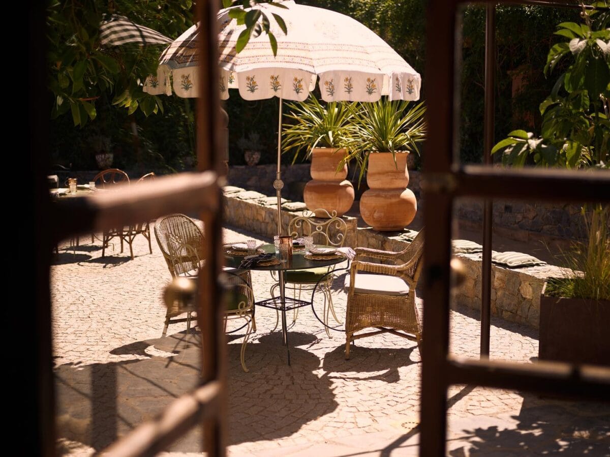 Une terrasse extérieure avec des chaises en osier, une petite table sous un grand parasol décoratif et deux plantes en pot est visible à travers une vitre. La scène, qui rappelle un lieu de rassemblement serein d'un club social, comprend des pavés et de la verdure.