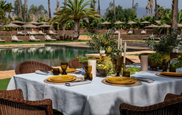 Une table dressée avec de la vaisselle, des verres et des bougies est placée au bord de la piscine avec des chaises en osier. En arrière-plan, des chaises longues et des parasols extérieurs sont visibles entourés de palmiers, évoquant une ambiance tranquille de Marrakech.