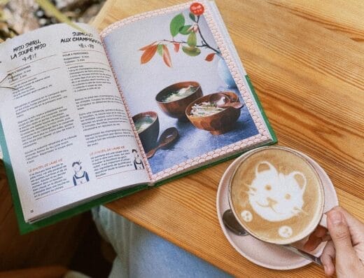 Personne tenant une tasse de café avec un motif d'ours dans la mousse, assise à une table en bois avec un livre de cuisine ouvert contenant une recette de soupe, évoquant un moment ukiyo serein.