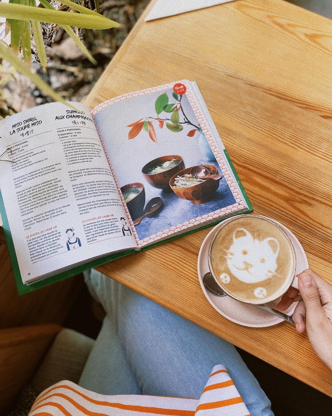 Personne tenant une tasse de café avec un motif d'ours dans la mousse, assise à une table en bois avec un livre de cuisine ouvert contenant une recette de soupe, évoquant un moment ukiyo serein.