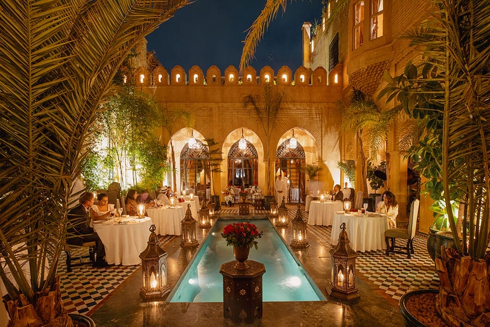 Une cour de Marrakech faiblement éclairée avec une piscine centrale, entourée de tables à manger éclairées aux bougies, d'arches complexes et d'une verdure luxuriante, parfaite pour savourer des moments inoubliables dans l'un des restaurants enchanteurs de la ville.