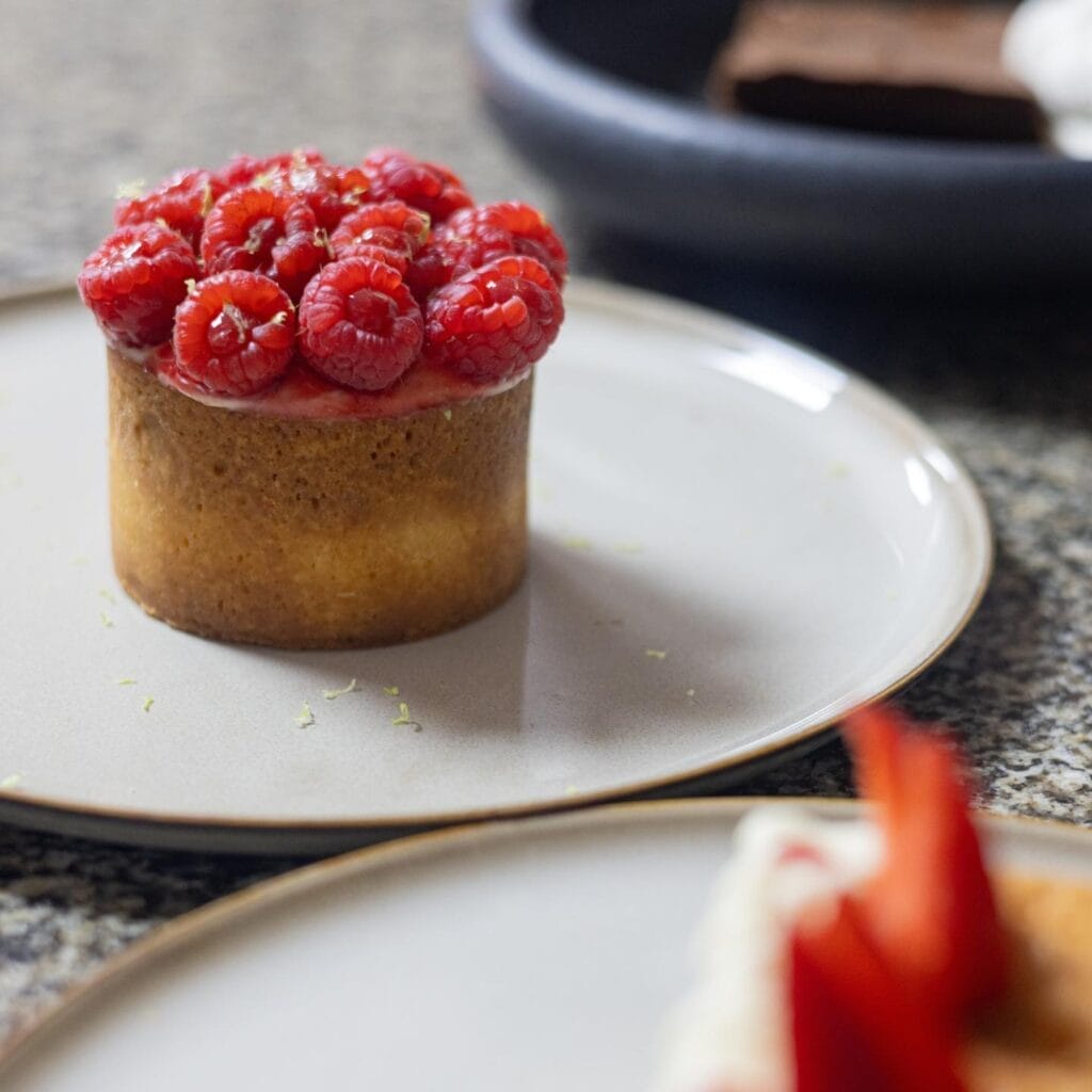 Une pâtisserie garnie de framboises fraîches trône élégamment sur une assiette blanche, incarnant l'essence du moment. Un autre dessert est flou en arrière-plan, laissant deviner un joyau caché que l'on trouve dans les meilleurs restaurants de Marrakech.