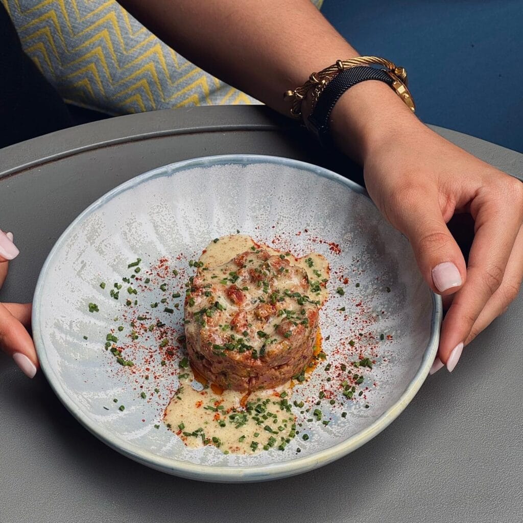 Une personne aux ongles manucurés tient une assiette comportant un plat rond garni d'herbes et de sauce, posé sur une table grise - un véritable délice culinaire du moment inspiré des saveurs des meilleurs restaurants de Marrakech.