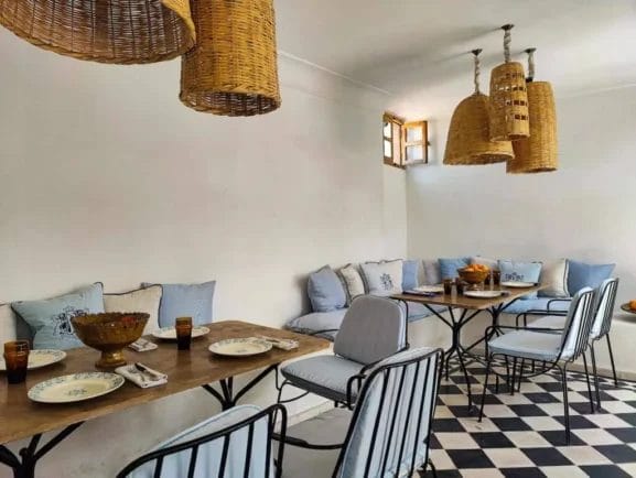 Vivez un moment Marrakech dans cette salle à manger cosy avec suspensions en osier, coussins bleus sur bancs, chaises en métal, tables en bois et carrelage à carreaux noir et blanc.