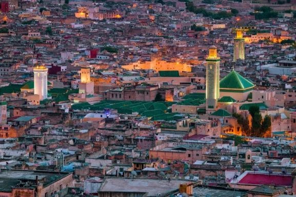 Vue aérienne d'un paysage urbain densément peuplé avec des mosquées et des toits illuminés au crépuscule, capturant le charme enchanteur de la vieille Fès, l'une des célèbres villes impériales du Maroc.