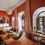 Une chambre chaleureusement éclairée dans un riad Marrakech aux murs rouges, comprenant un lit, un coin salon, une petite table et un luminaire décoratif. Deux grandes fenêtres avec rideaux apportent de la lumière naturelle pour votre séjour Marrakech parfait.