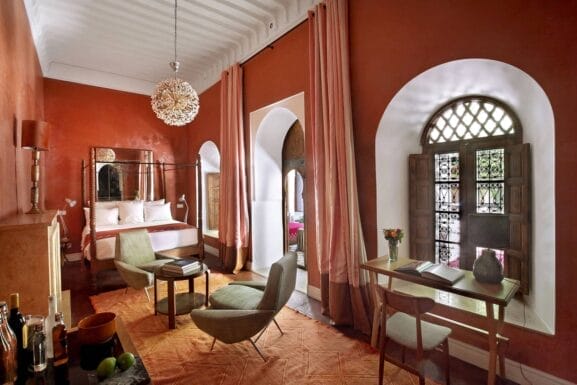 Une chambre chaleureusement éclairée dans un riad Marrakech aux murs rouges, comprenant un lit, un coin salon, une petite table et un luminaire décoratif. Deux grandes fenêtres avec rideaux apportent de la lumière naturelle pour votre séjour Marrakech parfait.