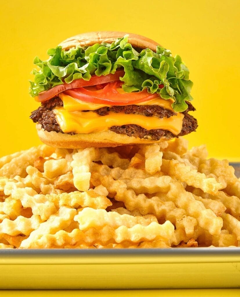 Un cheeseburger garni de laitue, de tomates et d'oignons est posé sur une grosse pile de frites ondulées sur un fond jaune, capturant un moment parfait rappelant les restaurants animés de Marrakech.