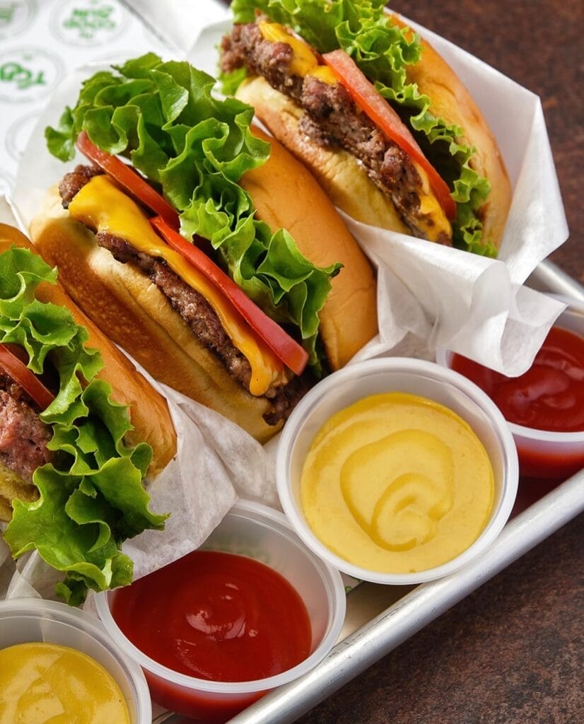 Trois hamburgers avec de la laitue, des tomates et du fromage sont disposés sur un plateau comme un festin de Marrakech, accompagnés de tasses de ketchup et de moutarde, rappelant les restos animés.